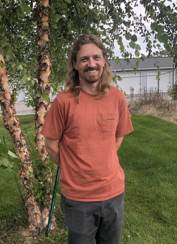 Reqan in a rust colored T shirt standing by a river birch smiling.